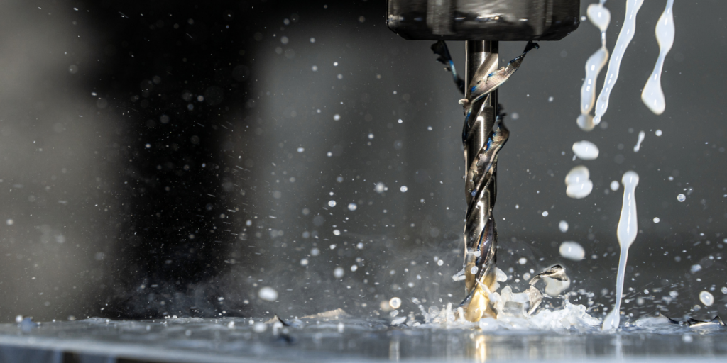 Ein Spiralbohrer fräst/bohrt in ein Metallwerkstück, während Kühlschmiermittel in dünnen Strahlen über das Werkzeug fließt und feine Späne sowie Flüssigkeitstropfen in die Luft spritzen; Nahaufnahme vor unscharfem, dunklem Hintergrund.
