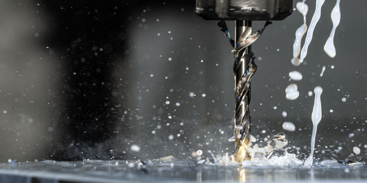 Ein Spiralbohrer fräst/bohrt in ein Metallwerkstück, während Kühlschmiermittel in dünnen Strahlen über das Werkzeug fließt und feine Späne sowie Flüssigkeitstropfen in die Luft spritzen; Nahaufnahme vor unscharfem, dunklem Hintergrund.