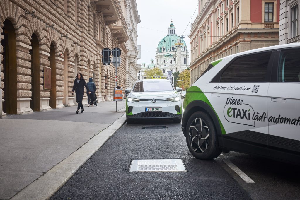 Weißes eTaxi mit Aufschrift ‚Dieses eTAXI lädt automatisch' steht auf einer induktiven Ladeplatte im Straßenpflaster einer Wiener Altstadtstraße, im Hintergrund die grüne Kuppel der Karlskirche.