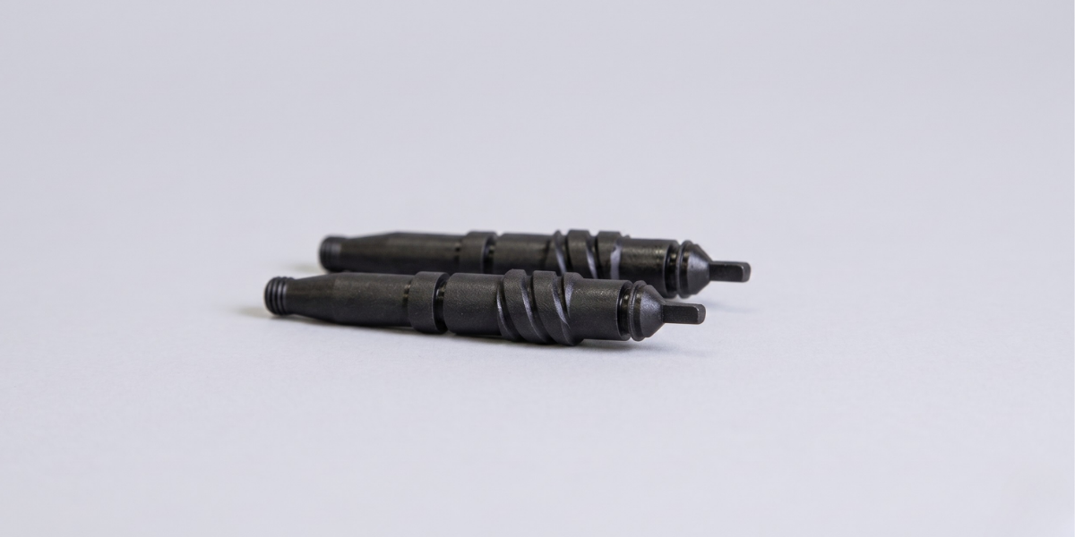 Dos componentes de precisión idénticos, de color negro mate, yacen paralelos uno detrás de otro sobre una superficie neutra de color gris claro. Los objetos son alargados y presentan un complejo diseño industrial fosfatado.