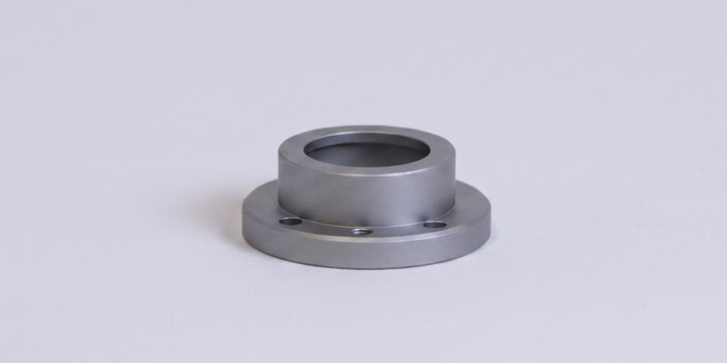 Hard-anodized aluminum turned part with a flange, a matte gray finish, and a visible internal bore, manufactured in accordance with DIN EN ISO 10074.