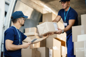 Young courier using touchpad while loading packages with his coworker in a delivery van.